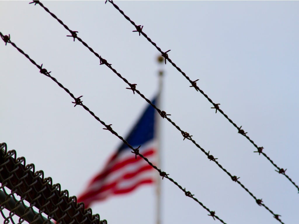 Barbed wire in the foreground with a blurred American flag in the background