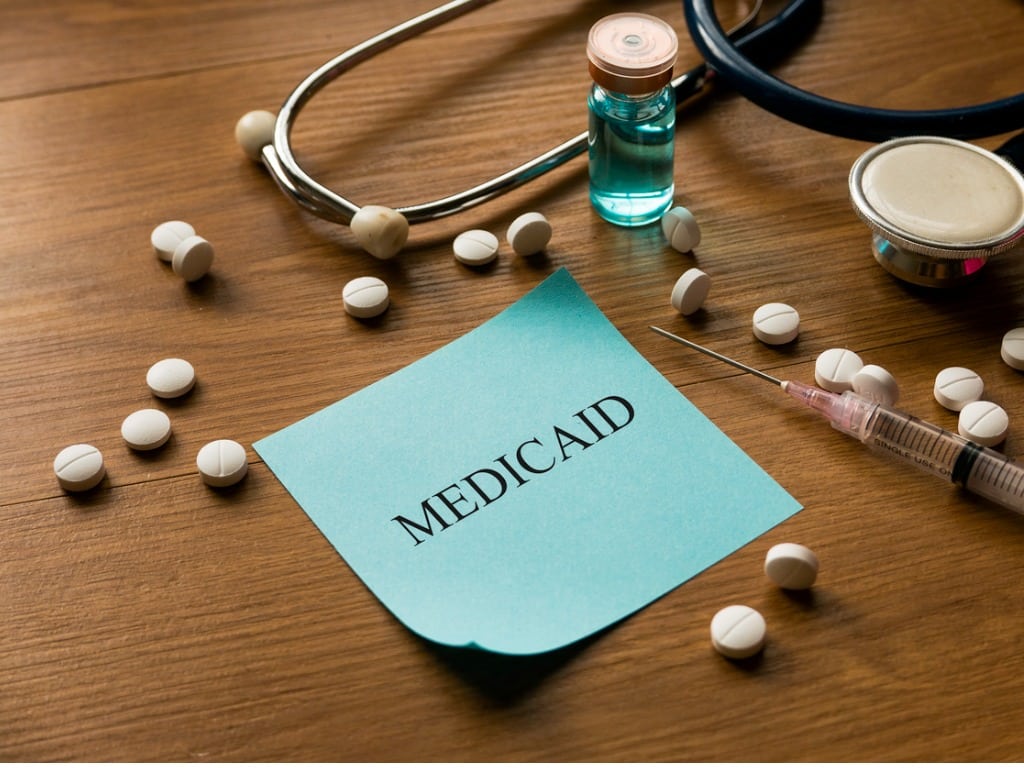 Sticky note that says "Medicaid" surrounded by pills and a syringe on a wooden table